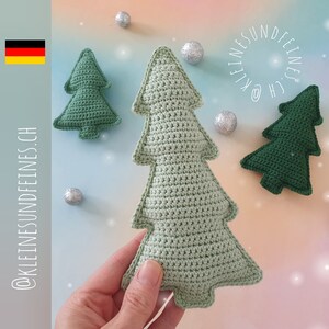 Könnte beinhalten: Zwei gehäkelte Weihnachtsbaum-Anhänger, einer grün und einer hellgrün, vor einem weißen Hintergrund. Der größere Anhänger wird in einer Hand gehalten. Der Text "@KLEINESUNDFEINES.CH" befindet sich in der linken unteren Ecke des Bildes. Der Text "KLEINES UND FEINES" befindet sich in einem Kreis in der rechten oberen Ecke des Bildes.
