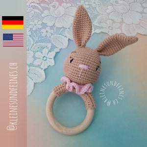 Könnte beinhalten: Ein gehäkeltes Hasenrassel mit rosa Akzenten und einem Holzring. Der Hase hat gestickte Augen und eine rosa Rüschen um den Hals. Die Rassel ist ein tolles Spielzeug für Babys.