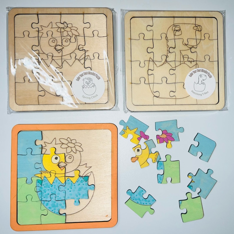 there are 2 wooden puzzles on a white background at the top Each one is just the outline -ready to be colored in. A baby chick hatching out of an egg with a flower on its head and a baby t-rex hatching out of an egg. Both are in cellophane a packaging sticker that says "Color your own wooden puzzle!" Below the packaged puzzles is a chick puzzle that has already been colored in bright yellows, blues and greens. Half of the puzzle is put together the other is spread across the photo.