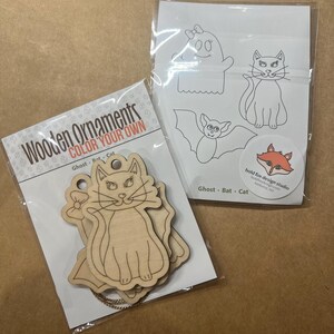 May include: Two packages of wooden ornaments and coloring pages. One package contains wooden cat ornaments with the text "Wooden Ornaments COLOR YOUR OWN". The other package has line drawings of a ghost, bat, and cat. A sticker with a fox logo is also visible.