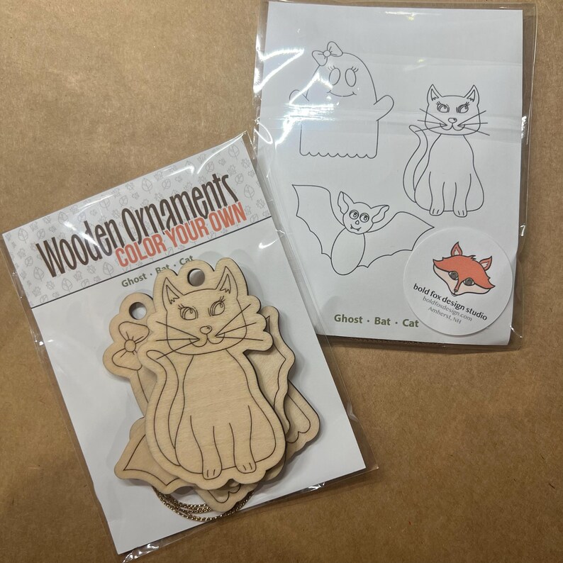 May include: Two packages of wooden ornaments and coloring pages. One package contains wooden cat ornaments with the text "Wooden Ornaments COLOR YOUR OWN". The other package has line drawings of a ghost, bat, and cat. A sticker with a fox logo is also visible.