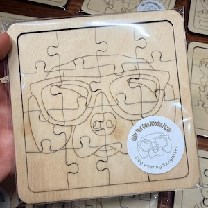 May include: A square wooden puzzle in clear plastic packaging featuring a laser-engraved outline of a dog wearing sunglasses. A circular white sticker on the bottom right displays the text Color Your Own Wooden Puzzle Dog wearing Sunglasses.