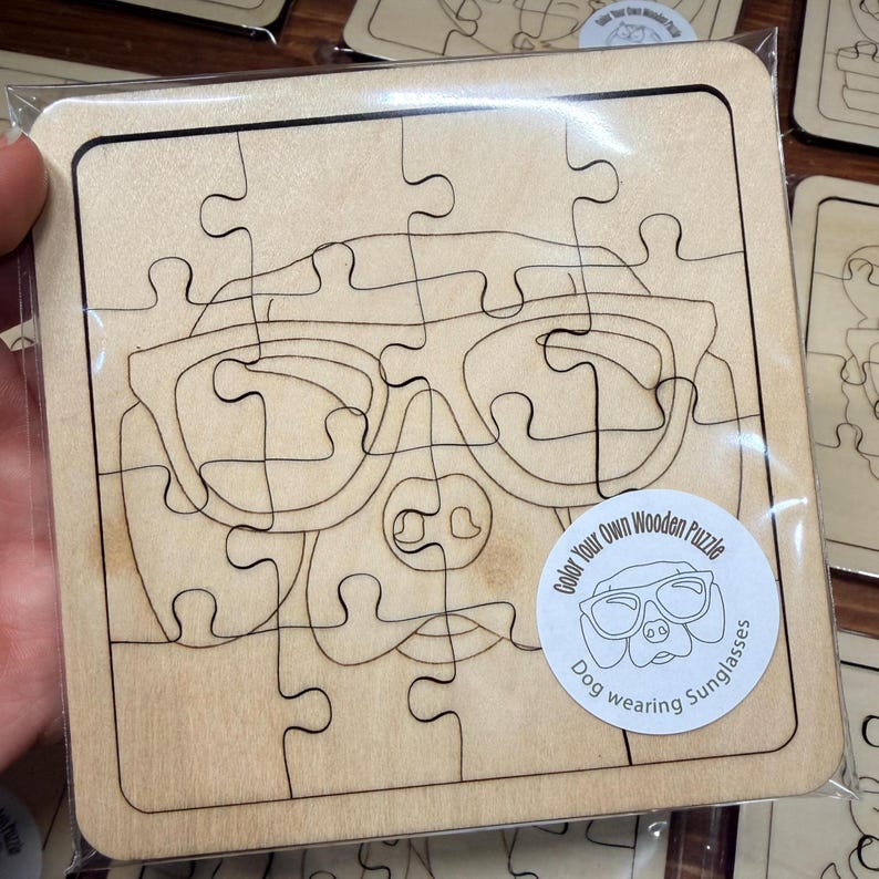 May include: A square wooden puzzle in clear plastic packaging featuring a laser-engraved outline of a dog wearing sunglasses. A circular white sticker on the bottom right displays the text Color Your Own Wooden Puzzle Dog wearing Sunglasses.