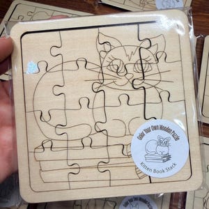 May include: A square wooden jigsaw puzzle featuring a line art illustration of a kitten sitting on a stack of books. The puzzle is sealed in clear plastic packaging and includes a circular sticker labelled Color Your Own Wooden Puzzle Kitten Book Stack.