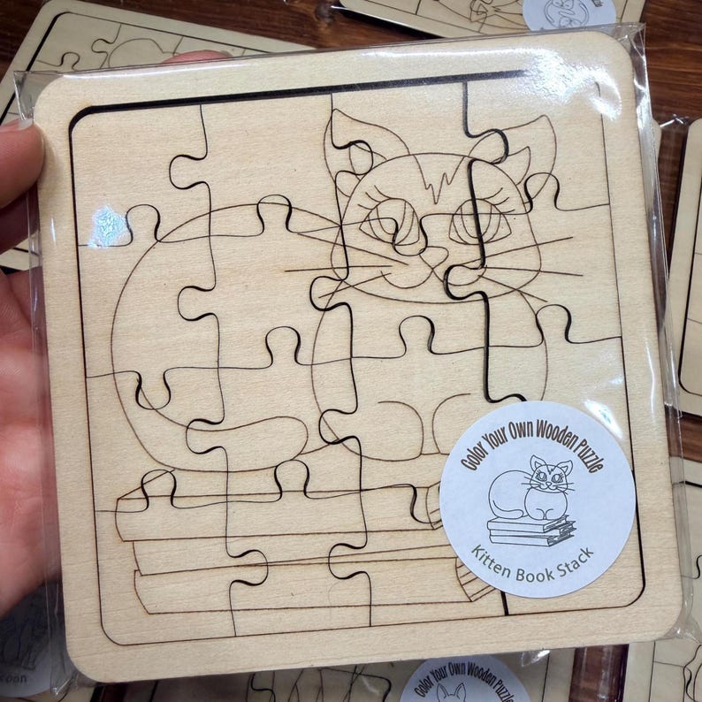May include: A square wooden jigsaw puzzle featuring a line art illustration of a kitten sitting on a stack of books. The puzzle is sealed in clear plastic packaging and includes a circular sticker labelled Color Your Own Wooden Puzzle Kitten Book Stack.