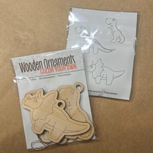 May include: Two packages of wooden dinosaur ornaments. One package contains three unfinished wooden dinosaur shapes with a silver chain. The other package contains a sheet with dinosaur outlines for coloring. The text on the package reads "Wooden Ornaments COLOR YOUR OWN".