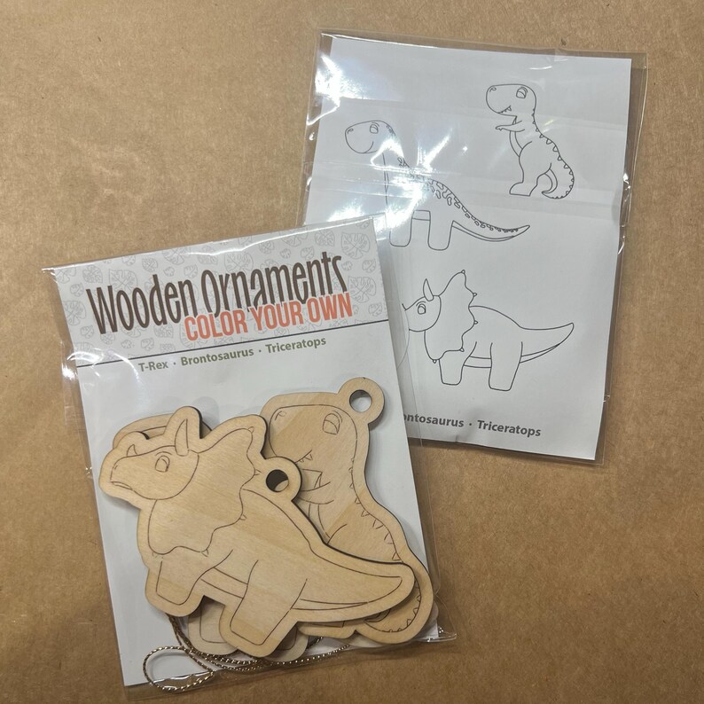 May include: Two packages of wooden dinosaur ornaments. One package contains three unfinished wooden dinosaur shapes with a silver chain. The other package contains a sheet with dinosaur outlines for coloring. The text on the package reads "Wooden Ornaments COLOR YOUR OWN".
