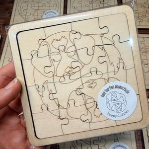 May include: A square wooden jigsaw puzzle featuring a laser-engraved line art illustration of two puppies cuddling. The puzzle is encased in clear plastic packaging and includes a circular sticker labelled Color Your Own Wooden Puzzle Puppy Cuddles.