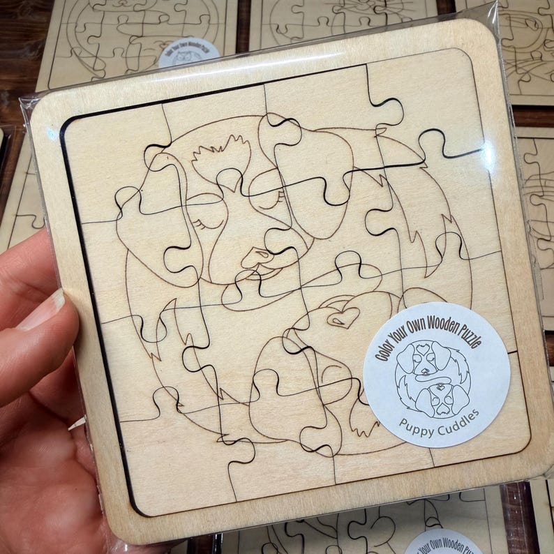 May include: A square wooden jigsaw puzzle featuring a laser-engraved line art illustration of two puppies cuddling. The puzzle is encased in clear plastic packaging and includes a circular sticker labelled Color Your Own Wooden Puzzle Puppy Cuddles.