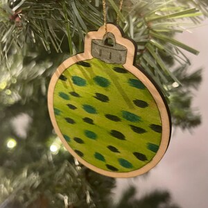 May include: Wooden Christmas ornament in the shape of a bauble. The ornament has a light green background with dark green and black spots. The ornament is trimmed with a light brown border and a gold string for hanging.