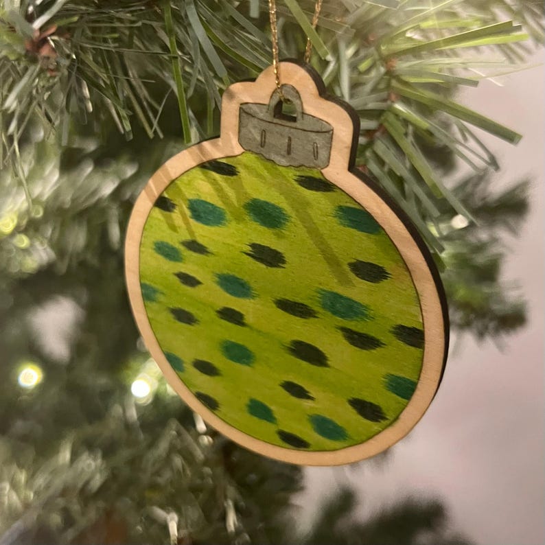 May include: Wooden Christmas ornament in the shape of a bauble. The ornament has a light green background with dark green and black spots. The ornament is trimmed with a light brown border and a gold string for hanging.