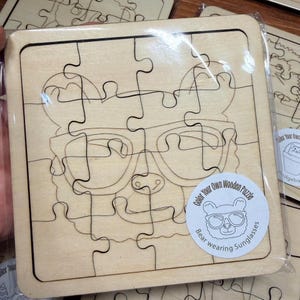 May include: A square wooden jigsaw puzzle featuring a laser-engraved outline of a bear wearing sunglasses. The puzzle is sealed in clear plastic packaging and includes a circular white sticker with the text Color Your Own Wooden Puzzle Bear wearing Sunglasses.