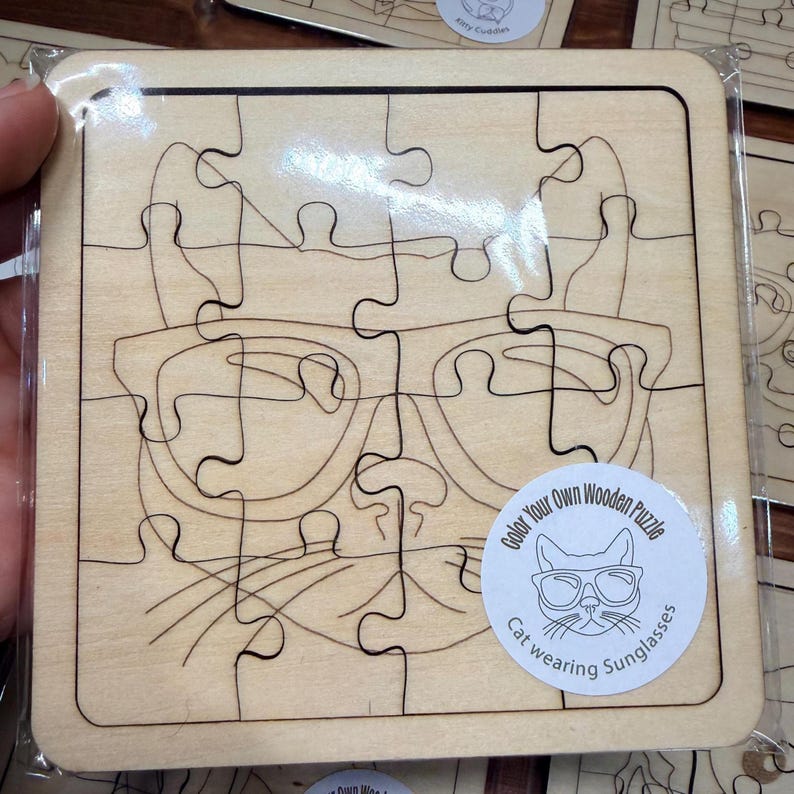May include: A square wooden jigsaw puzzle featuring a line art illustration of a cat wearing sunglasses. The puzzle is encased in clear plastic packaging and includes a circular white sticker with the text Color Your Own Wooden Puzzle Cat wearing Sunglasses.