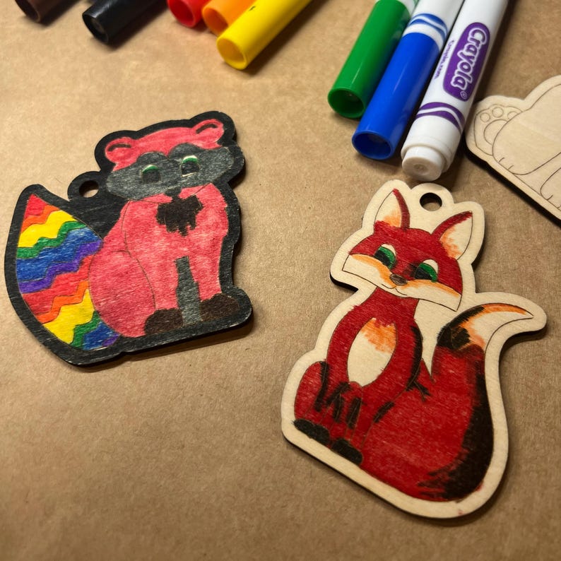 May include: Two wooden animal cutouts, a raccoon with a rainbow tail and a fox, are colored with markers. The raccoon is pink and black, and the fox is red and white. Crayola markers in various colors are in the background.
