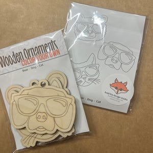 May include: Wooden ornaments for coloring, featuring a bear, dog, and cat wearing sunglasses. The package includes wooden cutouts and a sheet with line drawings of the same animals. The text "COLOR YOUR OWN" is visible.