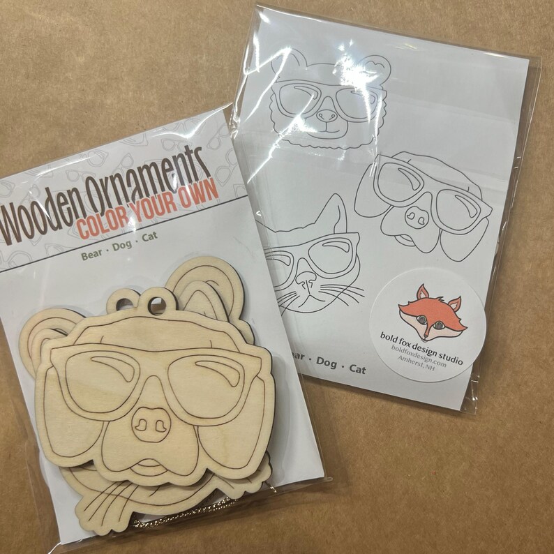 May include: Wooden ornaments for coloring, featuring a bear, dog, and cat wearing sunglasses. The package includes wooden cutouts and a sheet with line drawings of the same animals. The text "COLOR YOUR OWN" is visible.