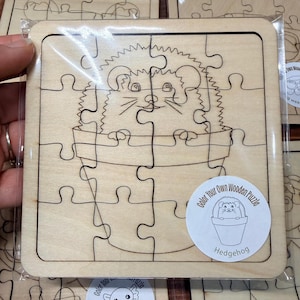 May include: A square wooden jigsaw puzzle featuring a laser-engraved illustration of a hedgehog sitting inside a flower pot. A circular white sticker on the bottom right corner displays the text Color Your Own Wooden Puzzle Hedgehog.