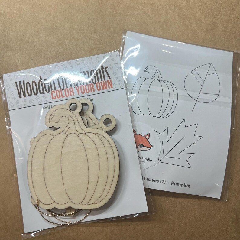 May include: Wooden pumpkin ornament with a string, packaged with a sheet of leaf and pumpkin outlines for coloring. The packaging reads "Wooden Ornaments COLOR YOUR OWN". A fox logo is also visible.