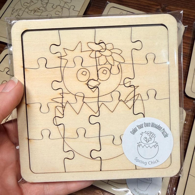 May include: A square wooden jigsaw puzzle featuring a line art illustration of a chick hatching from an egg with a flower on its head. A circular sticker on the bottom right corner displays the text Color Your Own Wooden Puzzle Spring Chick.