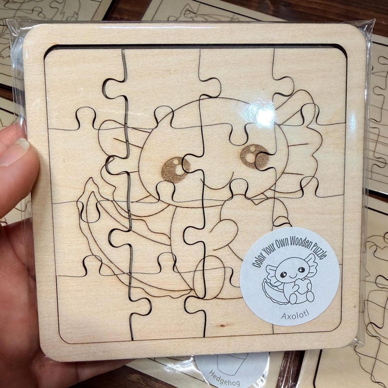 May include: A square wooden puzzle featuring a laser-engraved axolotl design. The puzzle consists of several interlocking pieces and includes a circular sticker in the bottom right corner with the text Color Your Own Wooden Puzzle and Axolotl.