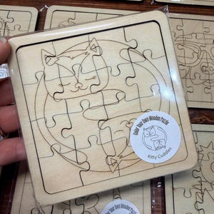 May include: A square wooden puzzle featuring a laser-engraved line drawing of two cats curled together in a circle. The puzzle pieces are interlocking and made of light-coloured wood. A circular sticker on the front reads Color Your Own Wooden Puzzle Kitty Cuddles.