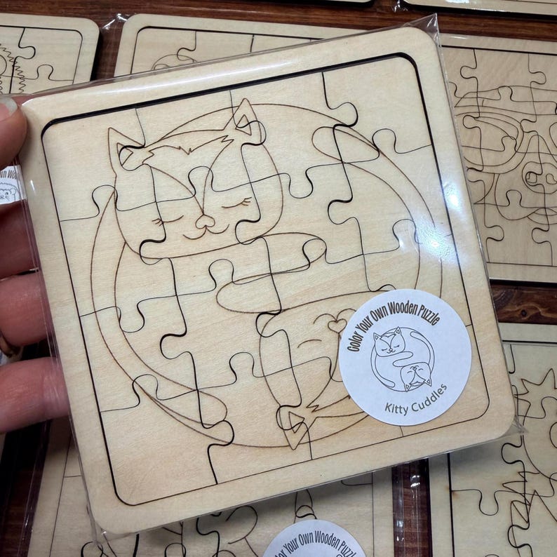 May include: A square wooden puzzle featuring a laser-engraved line drawing of two cats curled together in a circle. The puzzle pieces are interlocking and made of light-coloured wood. A circular sticker on the front reads Color Your Own Wooden Puzzle Kitty Cuddles.