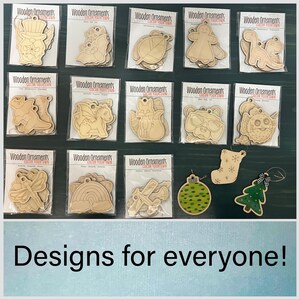 May include: A collection of wooden ornaments in various shapes, including a leaf, dinosaur, and gingerbread man. Some ornaments are packaged, while others are painted. The text "Designs for everyone!" is at the bottom.