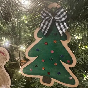 May include: Wooden Christmas tree ornament with a green painted tree shape, embellished with colorful gems. A black and white checkered bow is attached at the top. The ornament is hanging on a Christmas tree.