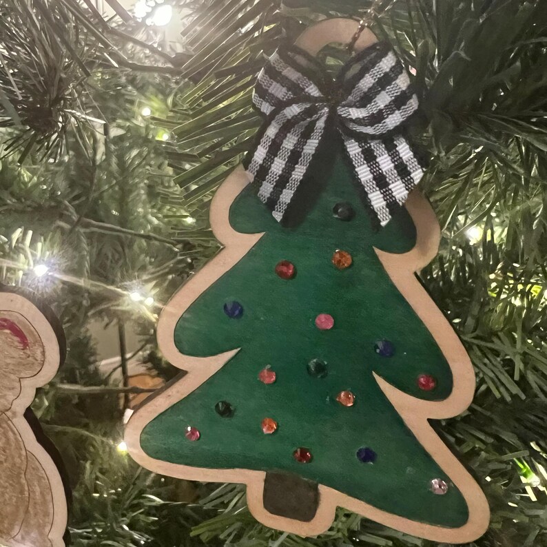 May include: Wooden Christmas tree ornament with a green painted tree shape, embellished with colorful gems. A black and white checkered bow is attached at the top. The ornament is hanging on a Christmas tree.