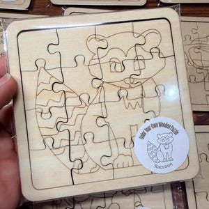 May include: A square wooden jigsaw puzzle featuring a laser-engraved raccoon outline. The puzzle is sealed in clear plastic packaging and includes a circular white sticker with the text Color Your Own Wooden Puzzle Raccoon and a small illustration.