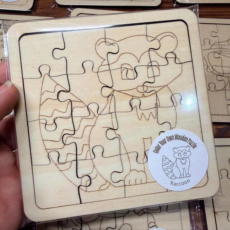 May include: A square wooden jigsaw puzzle featuring a laser-engraved raccoon outline. The puzzle is sealed in clear plastic packaging and includes a circular white sticker with the text Color Your Own Wooden Puzzle Raccoon and a small illustration.