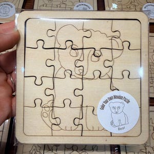 May include: A square wooden jigsaw puzzle featuring a line-art illustration of a bear. The puzzle is divided into twelve interlocking pieces and is enclosed in clear plastic packaging. A circular white sticker displays a bear and the text Color Your Own Wooden Puzzle Bear.
