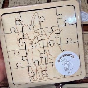 May include: A square wooden jigsaw puzzle featuring a line art illustration of a fox. The puzzle pieces are cut from light-coloured wood. A circular white sticker in the bottom right corner displays the text Color Your Own Wooden Puzzle Fox.