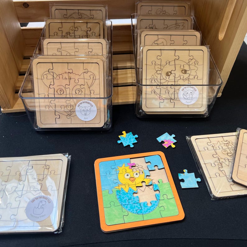 May include: Square wooden jigsaw puzzles featuring etched animal outlines, some packaged in clear plastic sleeves within transparent bins, and one partially coloured puzzle showing a yellow chick hatching from a blue egg with an orange frame.