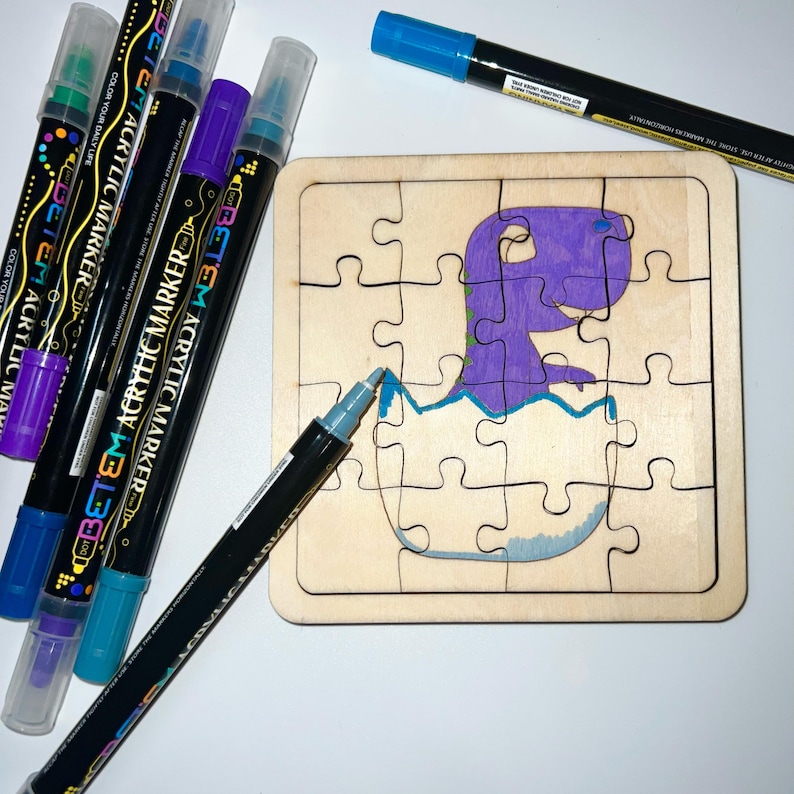 May include: A square wooden jigsaw puzzle featuring a purple dinosaur hatching from an egg, partially coloured with blue and purple acrylic markers. Several black acrylic markers with coloured caps are arranged around the puzzle on a white surface.