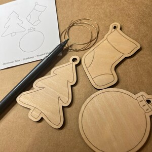 May include: Wooden Christmas ornaments in the shapes of a tree, stocking, and round ball. A black marker and gold string are next to the ornaments. A paper with the words "Christmas Tree - Stocking - Round Blu" is also in the image.