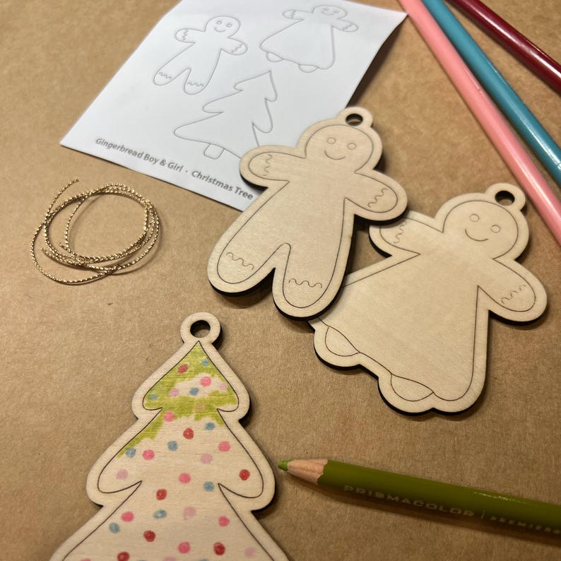 May include: Wooden gingerbread boy and girl ornaments and a Christmas tree ornament are shown, along with colored pencils, a gold string, and a paper with gingerbread and tree outlines. The tree ornament is decorated with pink, red, and blue dots.