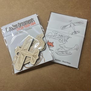 May include: Two packages of wooden airplane ornaments. One package contains unfinished wooden airplane cutouts with a gold chain. The other package has airplane outlines for coloring. Text on the package reads "Wooden Ornaments COLOR YOUR OWN".