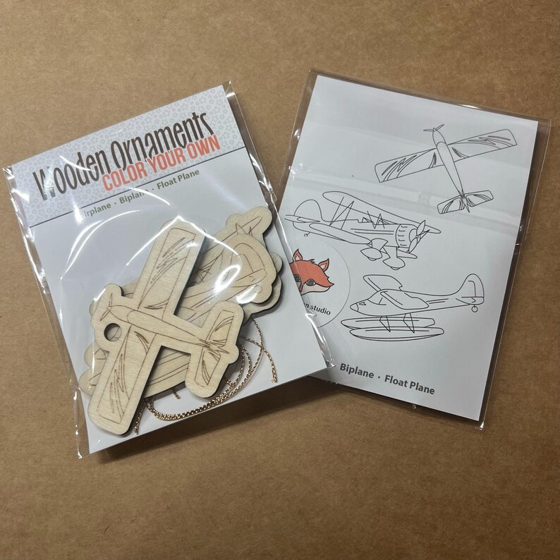 May include: Two packages of wooden airplane ornaments. One package contains unfinished wooden airplane cutouts with a gold chain. The other package has airplane outlines for coloring. Text on the package reads "Wooden Ornaments COLOR YOUR OWN".