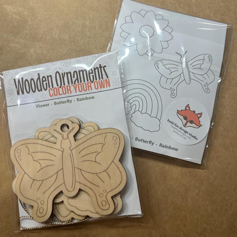 May include: Two packages of wooden ornaments. One package contains unfinished butterfly ornaments with a silver string. The other package has line drawings of a flower, butterfly, and rainbow. Text on the packages reads "Wooden Ornaments COLOR YOUR OWN Flower Butterfly Rainbow".