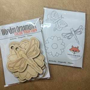 May include: Two packages of wooden ornaments. One package contains unfinished wooden butterfly, dragonfly, and flower shapes. The other package has line drawings of the same shapes. The text "Wooden Ornament, Color Your Own" is visible.