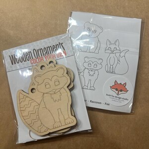 May include: Wooden ornaments with a raccoon design, ready to be colored. The package includes a set of wooden ornaments and a sheet with bear, raccoon, and fox outlines. The text "COLOR YOUR OWN" is visible on the packaging.
