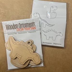 May include: Two packages of wooden ornaments. One package contains unfinished wooden dinosaur shapes with the text "Wooden Ornaments COLOR YOUR OWN". The other package contains dinosaur outlines for coloring. The dinosaurs include a hatching T-Rex, Brontosaurus, and Stegosaurus.