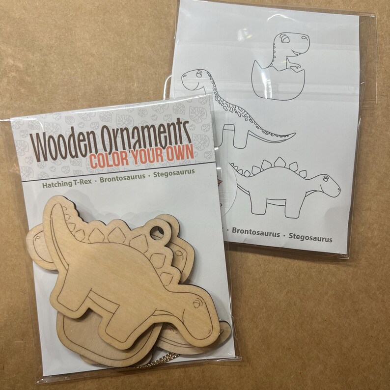 May include: Two packages of wooden ornaments. One package contains unfinished wooden dinosaur shapes with the text "Wooden Ornaments COLOR YOUR OWN". The other package contains dinosaur outlines for coloring. The dinosaurs include a hatching T-Rex, Brontosaurus, and Stegosaurus.