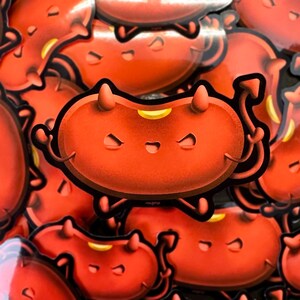 May include: A sticker featuring a cartoon devil bean character with a red body, small horns, and a curved tail. The character has a mischievous expression with squinting eyes and a small smile. The sticker has a black outline and the text "ITM@FUJI".