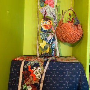 May include: A woven basket with a handle hangs from the ceiling, filled with dried leaves and berries. A long, colorful fabric garland hangs on the wall behind the basket, with a blue and white patterned tablecloth on a table below.