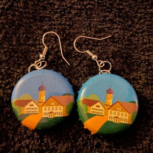 May include: A pair of earrings made from bottle caps with a painted image of a small town with a church and houses. The earrings are silver and have a hook closure.