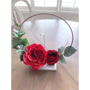 Puede incluir: Un soporte de anillo de madera blanca con un aro de metal y rosas rojas de seda. El soporte está decorado con vegetación.