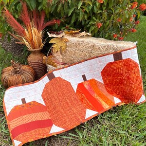 May include: A rectangular fabric table runner featuring a patchwork design of four stylized pumpkins in various shades of orange, red, and yellow. The pumpkins are set against a white background with a subtle, light-colored floral pattern.