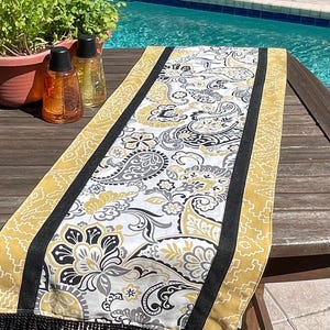 May include: A decorative table runner with a paisley pattern in white, black, and gold. The runner has a black border and gold fringe. The runner is placed on a wooden table near a pool.